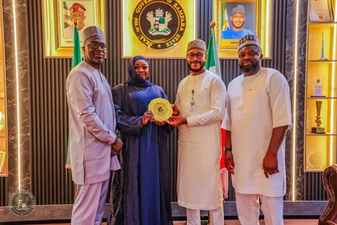 Four people in formal attire standing together indoors holding a golden award, with Nigerian flags and official portraits visible in the background