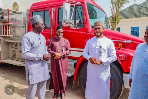 Three men in traditional attire standing beside a red emergency vehicle during an outdoor gathering